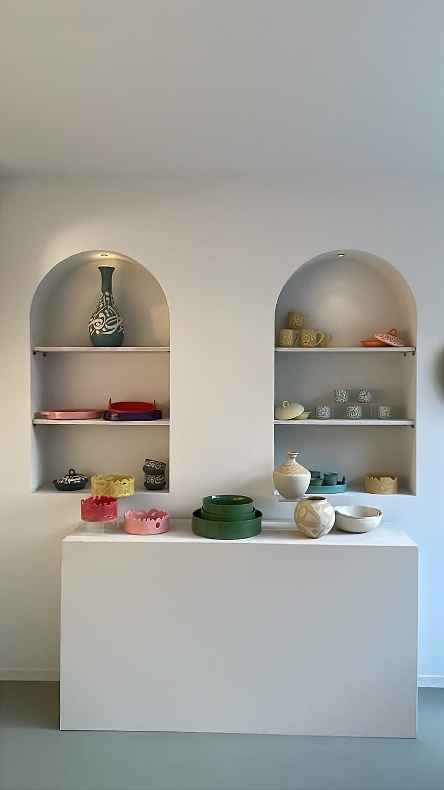 Two arched gallery niches displaying calligraphy vases, cups and colourful glazed bowls above a white plinth