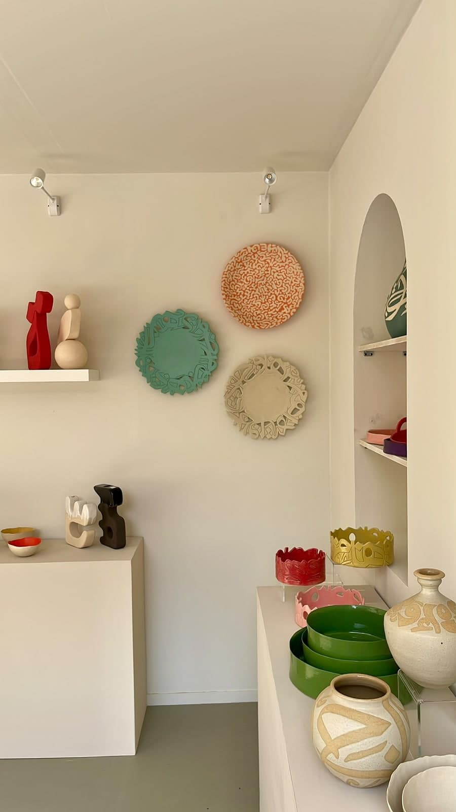 Gallery corner with calligraphy-cut ceramic wall plates, abstract sculptures on shelves, and glazed vessels and bowls on plinths