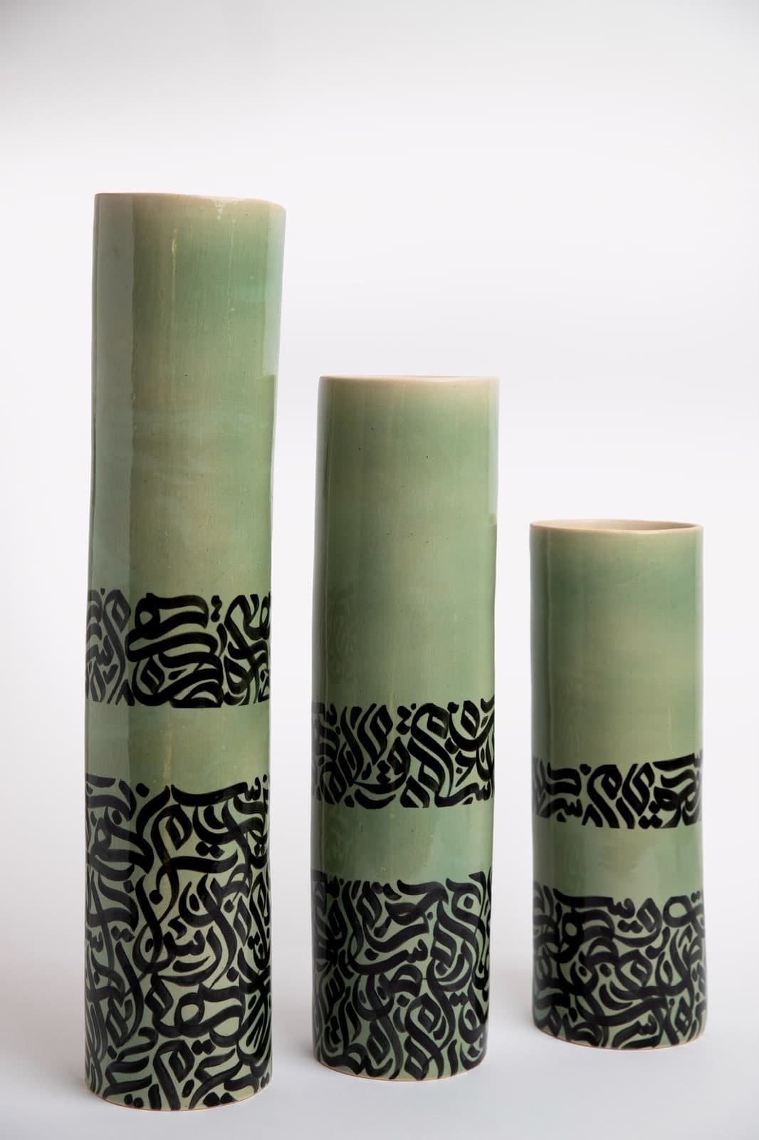 Three tall cylindrical ceramic vases in soft green glaze with bands of black Arabic calligraphy, standing in descending height
